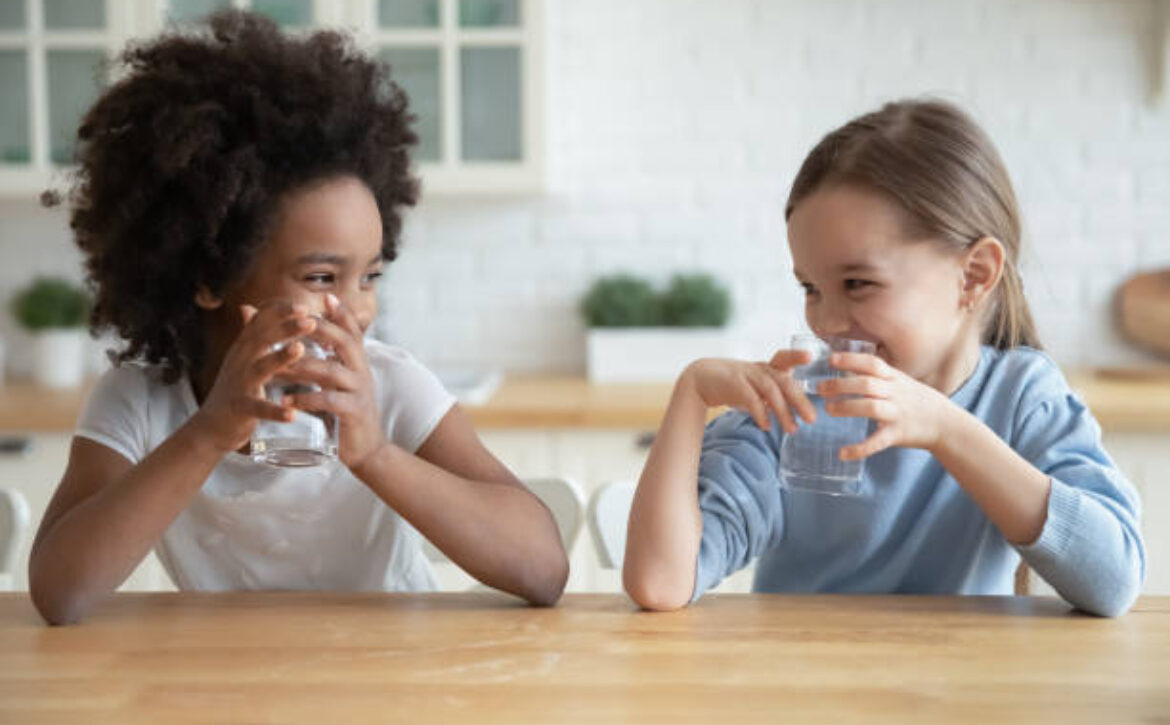 Two multiracial girls sit in kitchen drinking water